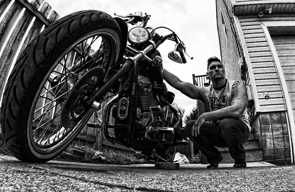 A Black and White Photo of a Man squatting next to his Harley Davidson Sportster